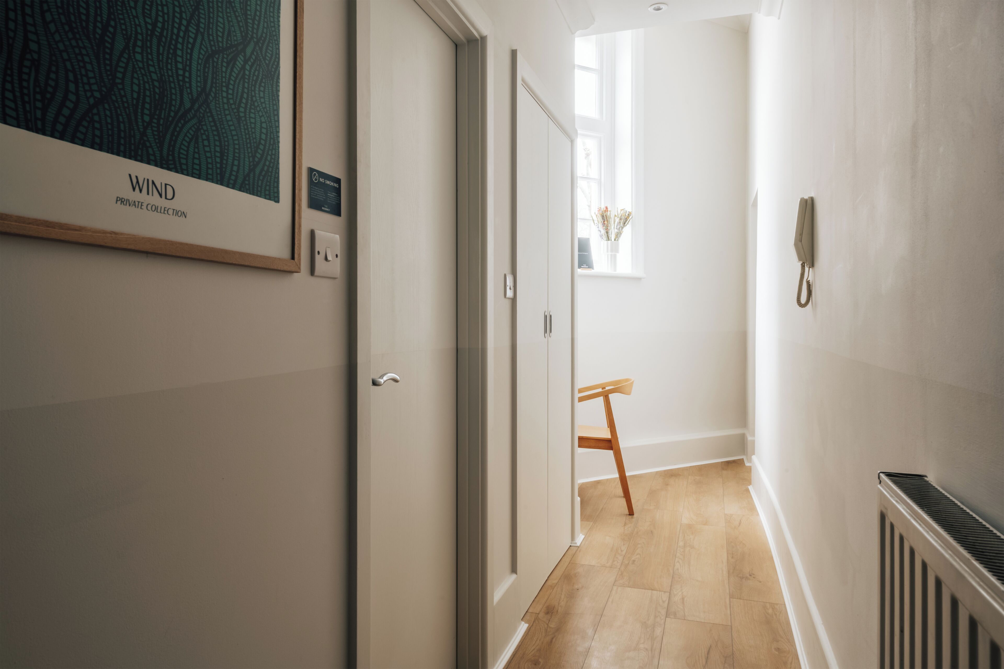 Un couloir orné d’une œuvre d’art encadrée intitulée « COLLECTION PRIVÉE – VENTS », avec une chaise en bois, un radiateur et une fenêtre laissant entrer la lumière naturelle.