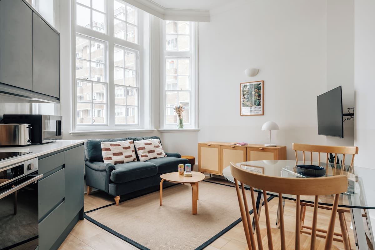 A modern kitchen with a built-in oven, a dining area with a glass table and wooden chairs, a sofa with patterned cushions, and large windows providing natural light.