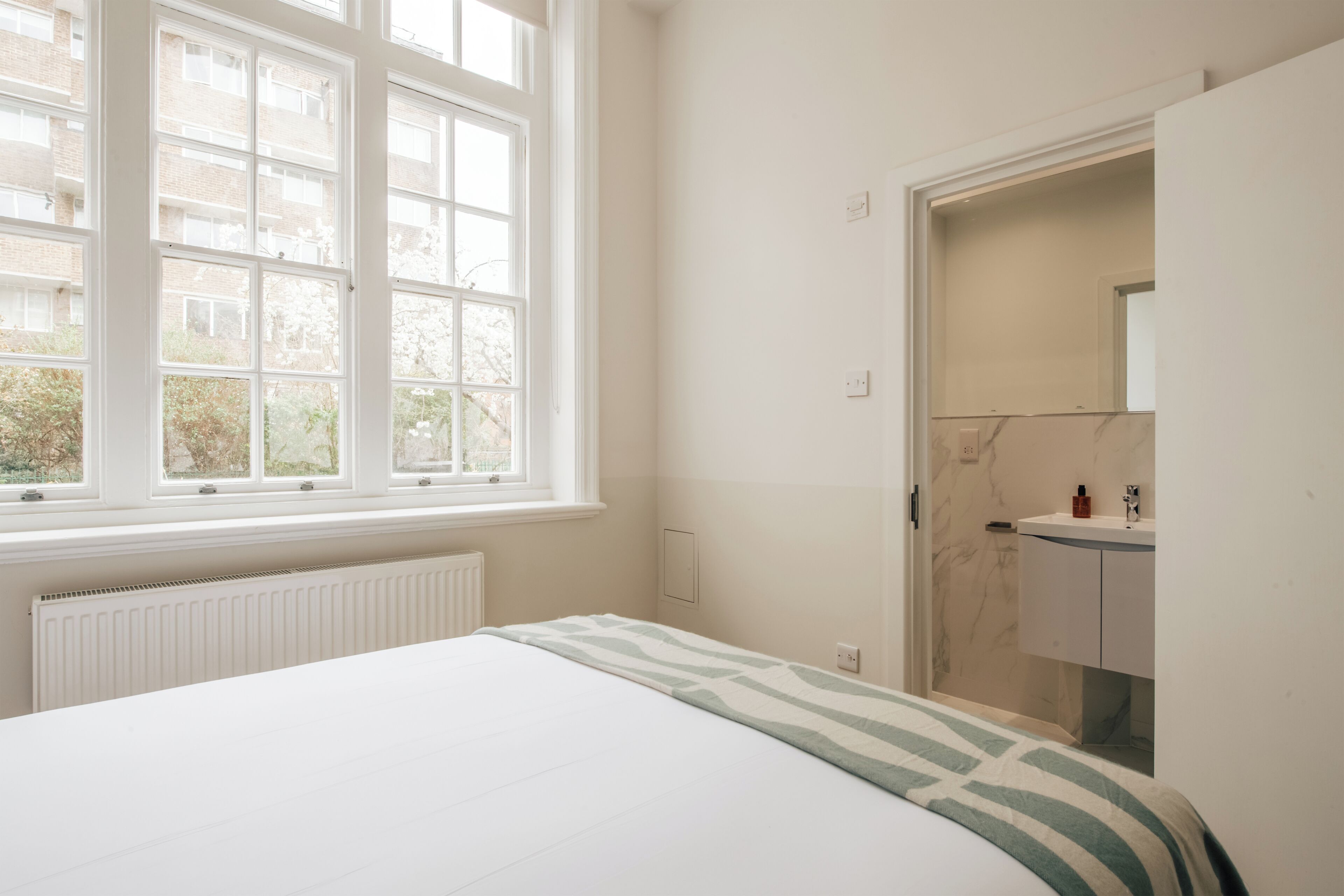 A bedroom with a large window, a bed, a radiator, and a bathroom with a marble wall and a sink.