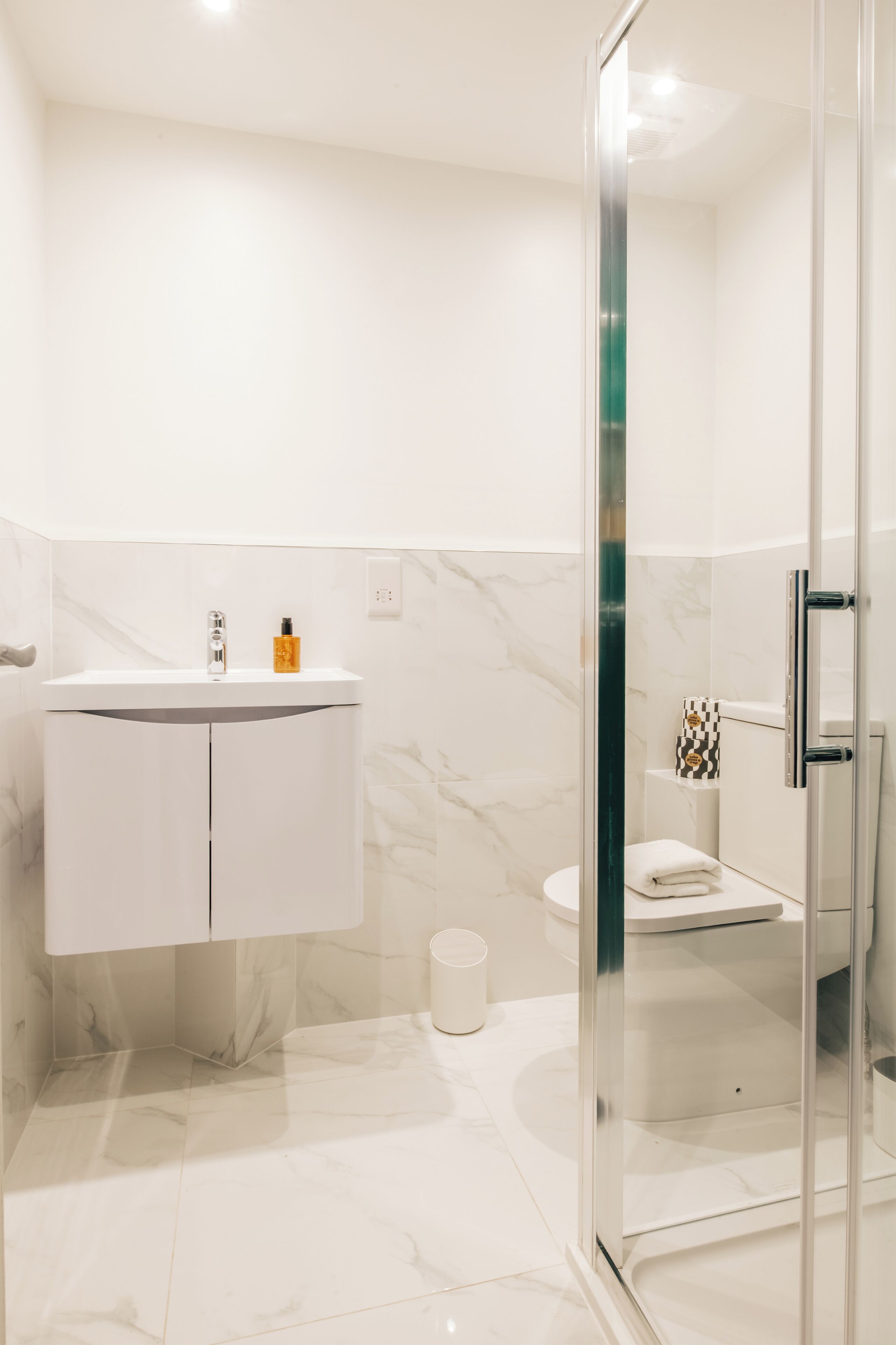 Une salle de bain moderne avec une douche à parois de verre, un lavabo blanc doté d’un plan de toilette en marbre et des toilettes agrémentées d’une serviette à carreaux.