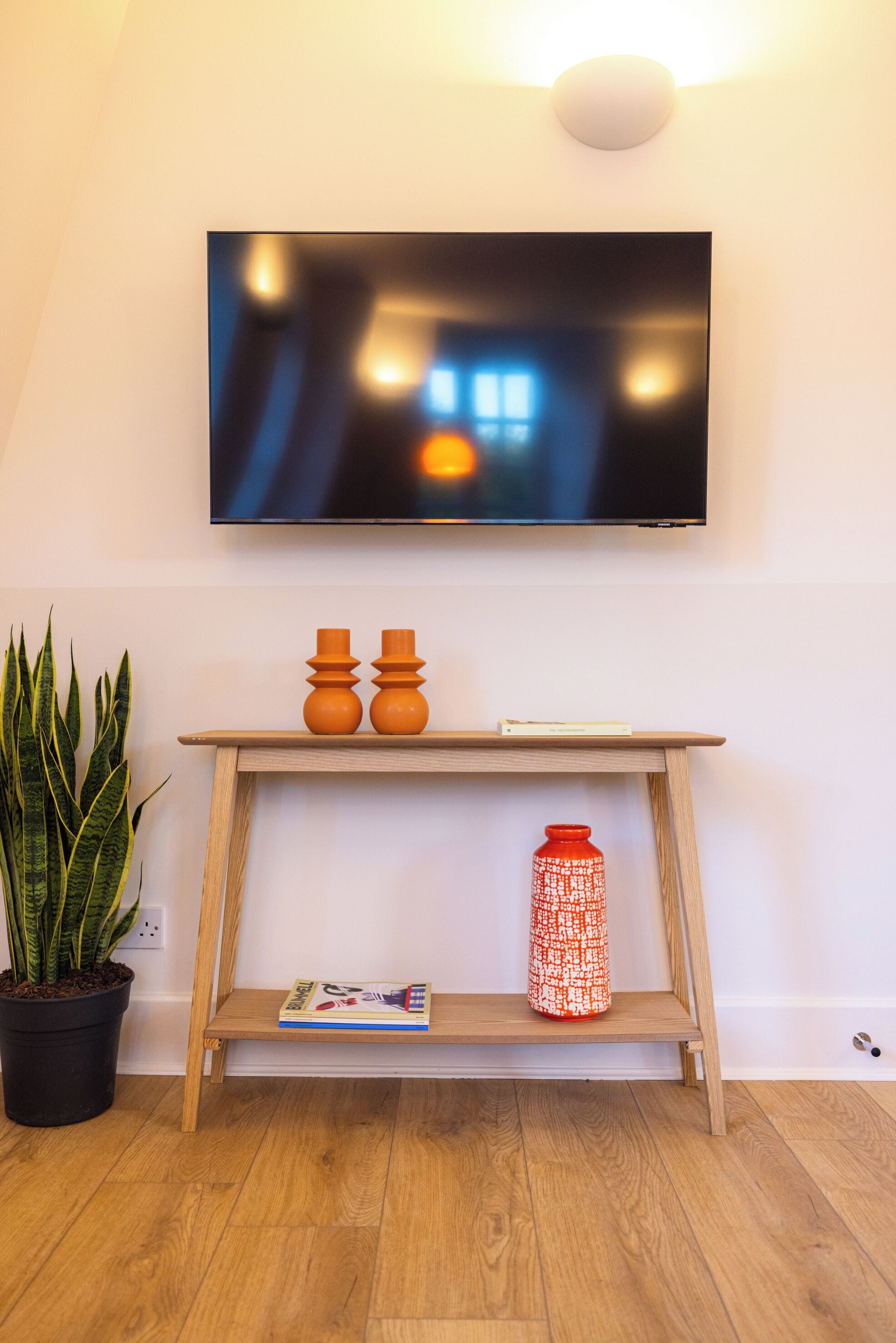 Et vægmonteret fladskærms-TV hænger over et konsolbord i træ med to orange vaser og en rød vase med mønster. En potteplante er placeret til venstre for bordet.