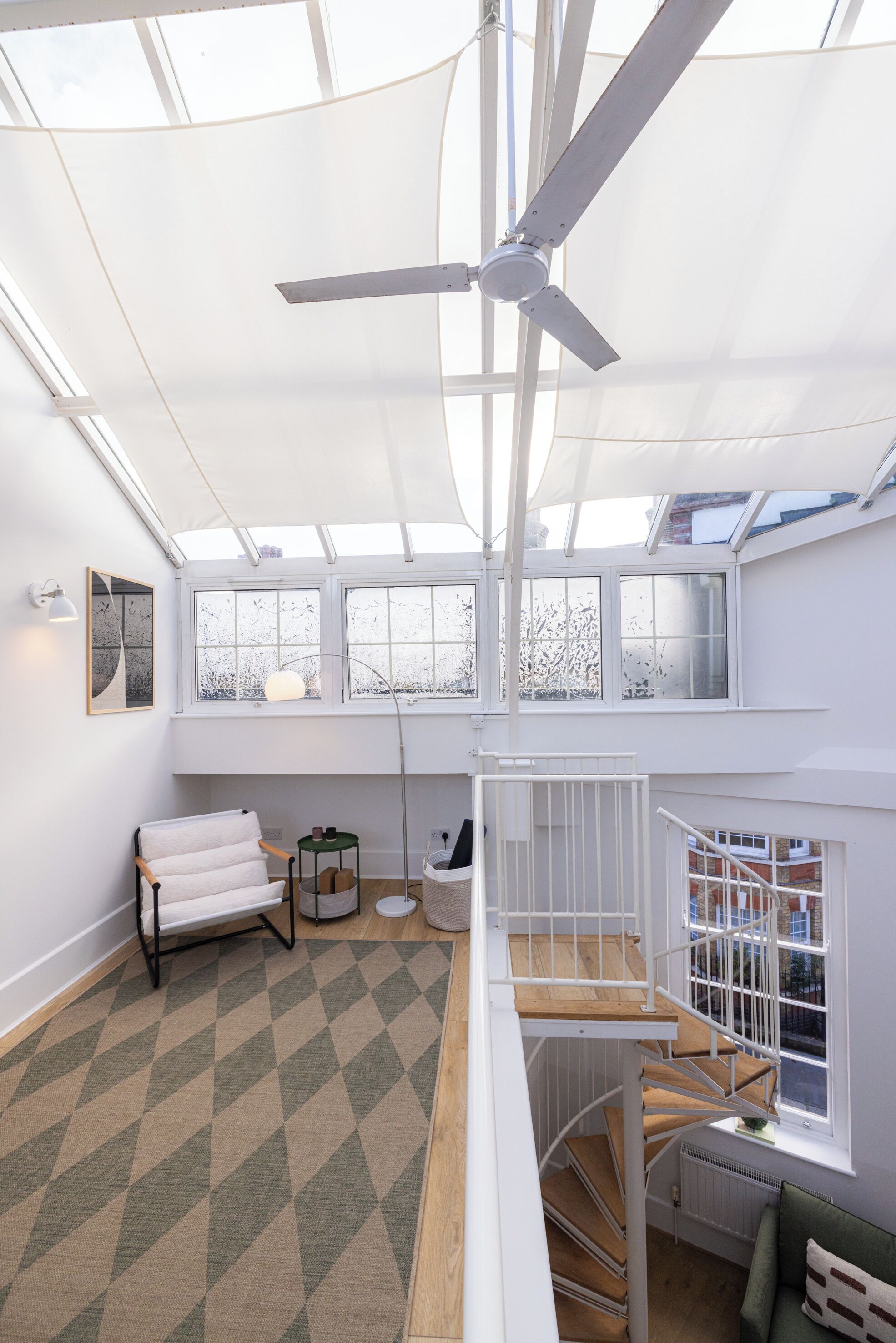 A spacious room with a high ceiling, a ceiling fan, a spiral staircase, and large windows providing natural light.
