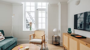 Ett modernt vardagsrum med en soffa, ett runt soffbord med snacks, en fåtölj i rotting, en tv på väggen och ett fönster med utsikt över en byggnad.