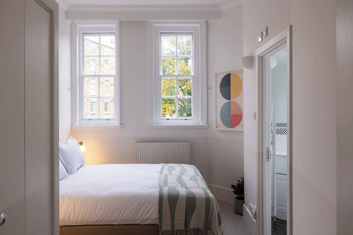 A bedroom with a bed, two windows, a radiator, a door, and a wall-mounted artwork.