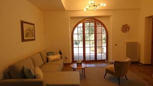 A living room with a sofa, armchair, and a coffee table. There is a large arched window with a view outside. - SCAPPO IN UMBRIA, Casale Fasulo (Umbria)