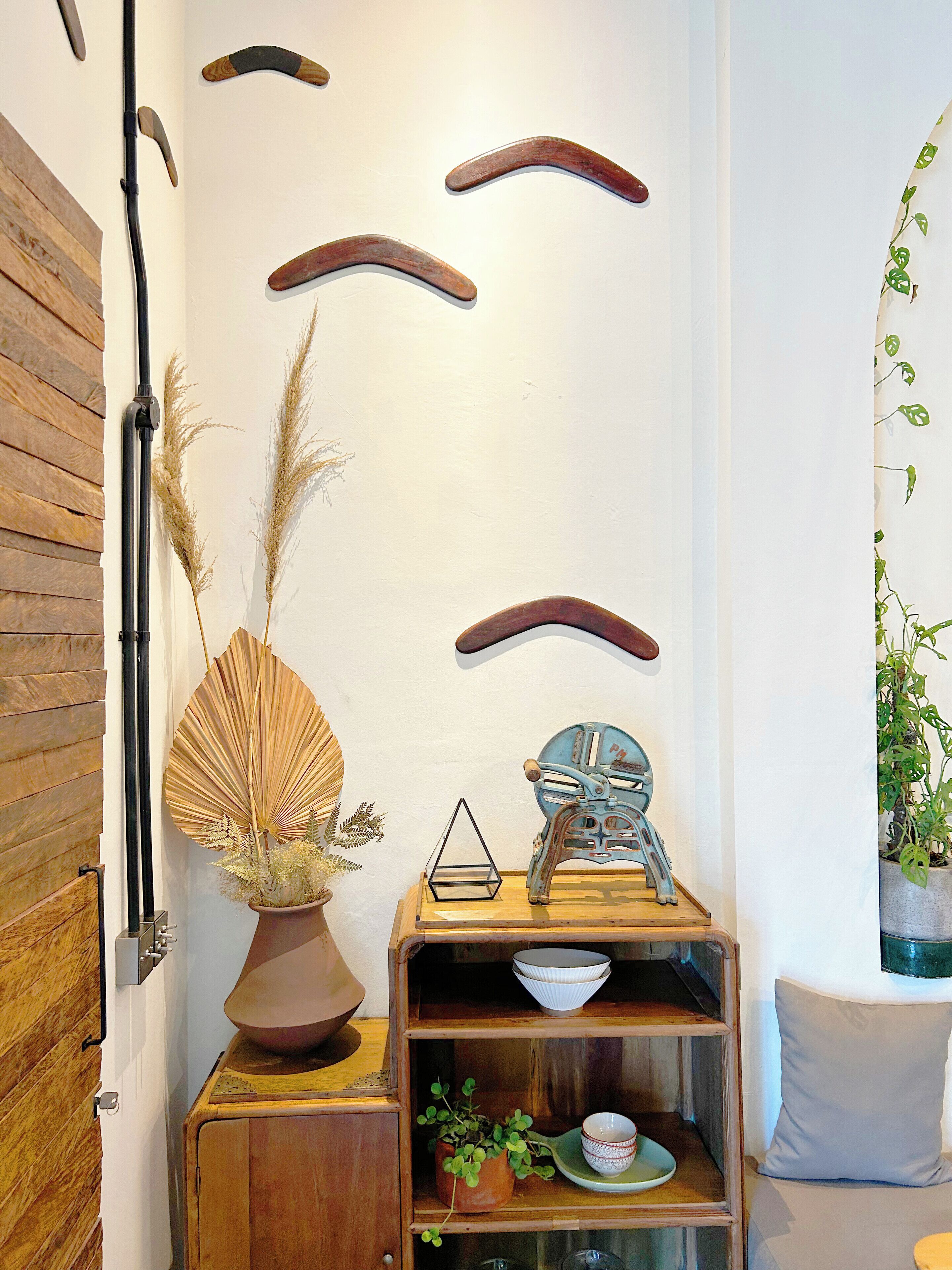 Un coin de pièce avec un meuble en bois, un vase contenant des plantes séchées, une sculpture en métal et une étagère fixée au mur, sur laquelle sont posés des bols.