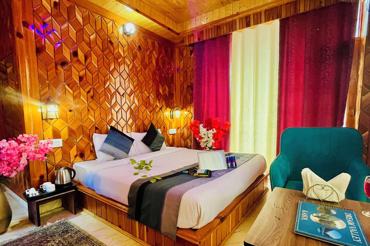 A hotel room with a wooden headboard, a bed with pillows, a bedside table with a lamp, a chair, and a small table with a book.