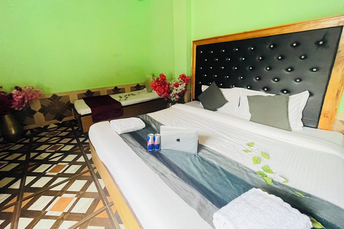 A hotel room with a bed, a desk, and a chair. There is a laptop on the bed, a vase with flowers, and a window with a view of the outside.