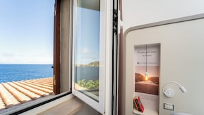 A room with a large window overlooking the sea, a wall-mounted light fixture, and a framed picture of a sunset. - Hotel Saraceno (Isola del Giglio)