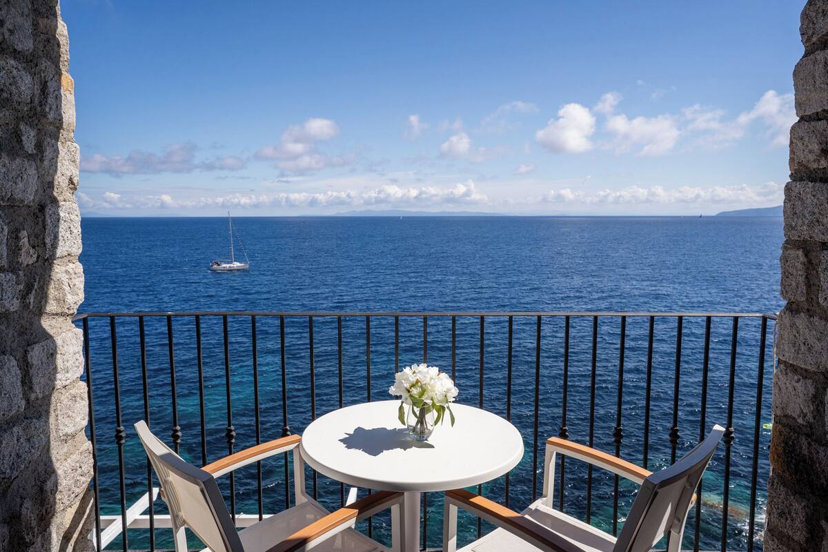 En balkon med et rundt bord og to stole, med udsigt over et klart, blåt hav med en sejlbåd i horisonten.