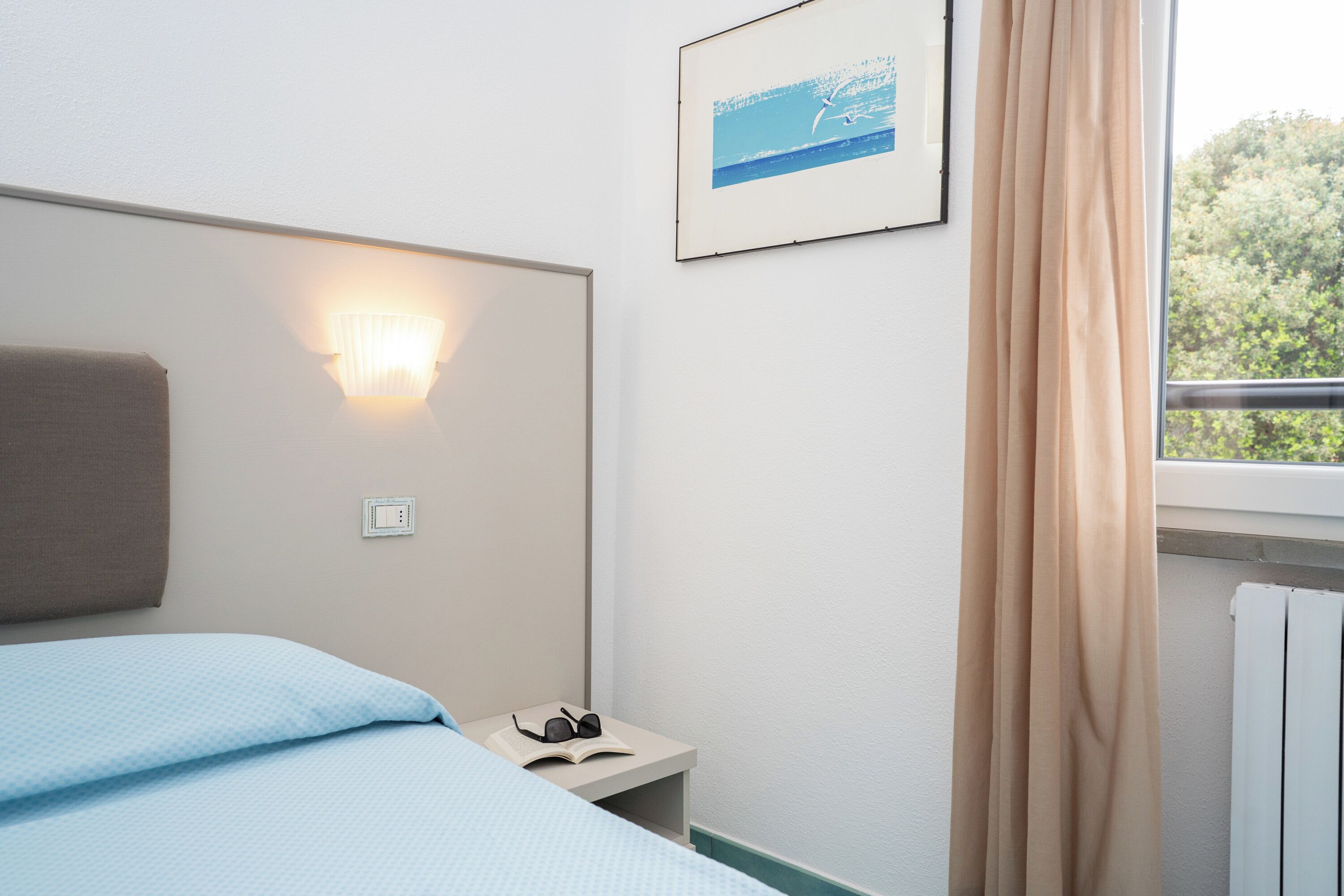 Habitación de hotel con cama, mesita de noche con lámpara, un cuadro enmarcado en la pared y una ventana con cortinas.