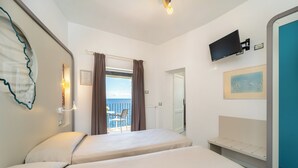 A hotel room with two beds, a TV mounted on the wall, a painting, and a view of the sea through the window. - Hotel Saraceno (Isola del Giglio)