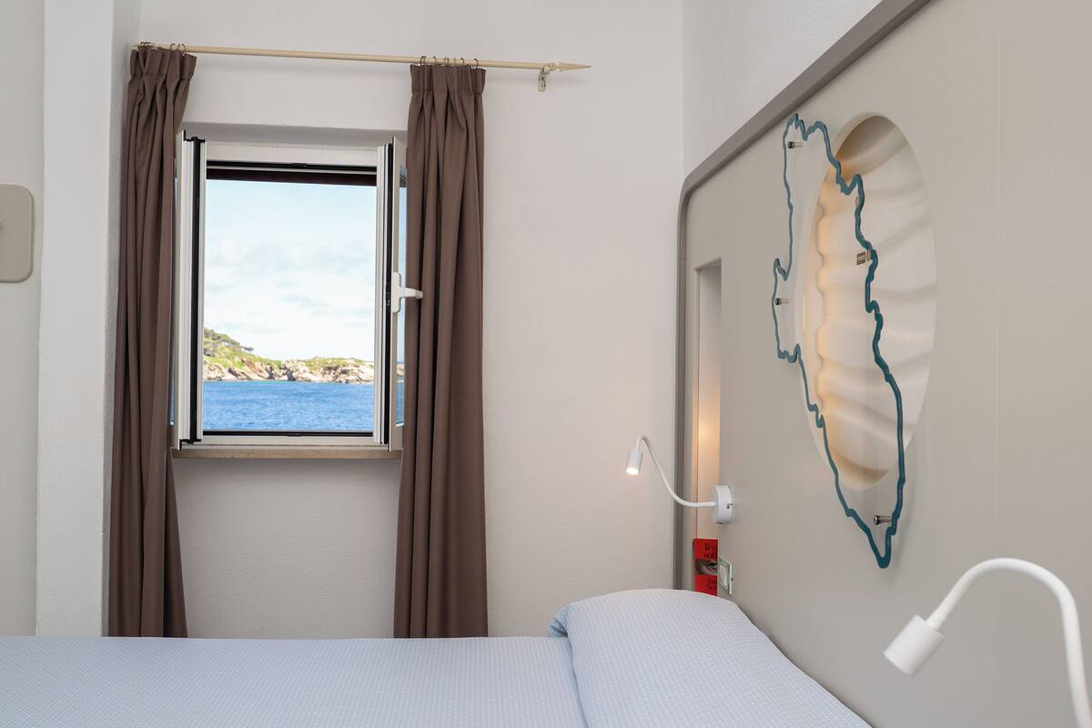 Et soveværelse med et vindue, gardiner, en seng, en lampe på væggen og en dekorativ lysarmatur.