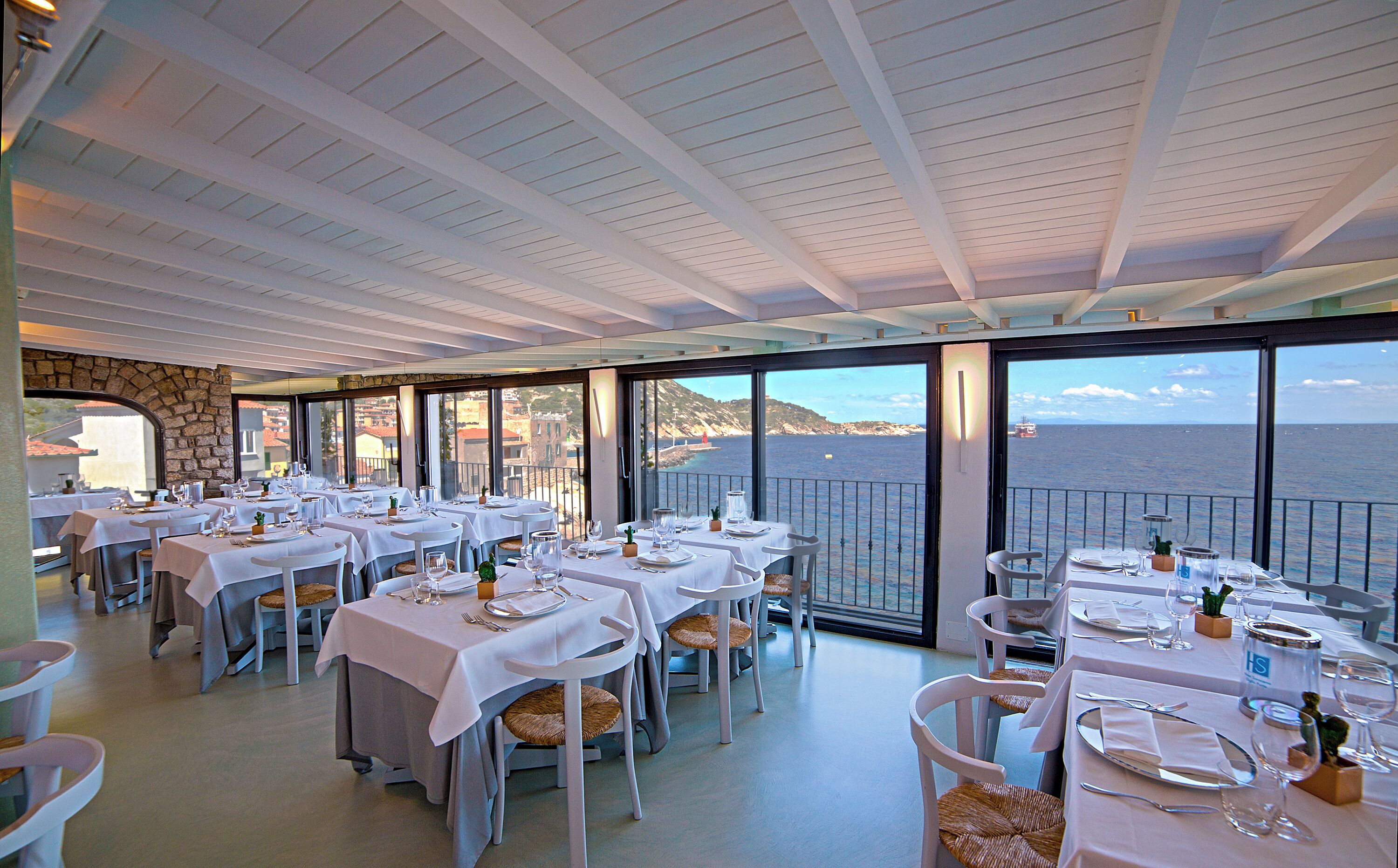 Un restaurante con mesas dispuestas para comer, amplias ventanas que ofrecen vista al mar y una pared de piedra en un lateral.