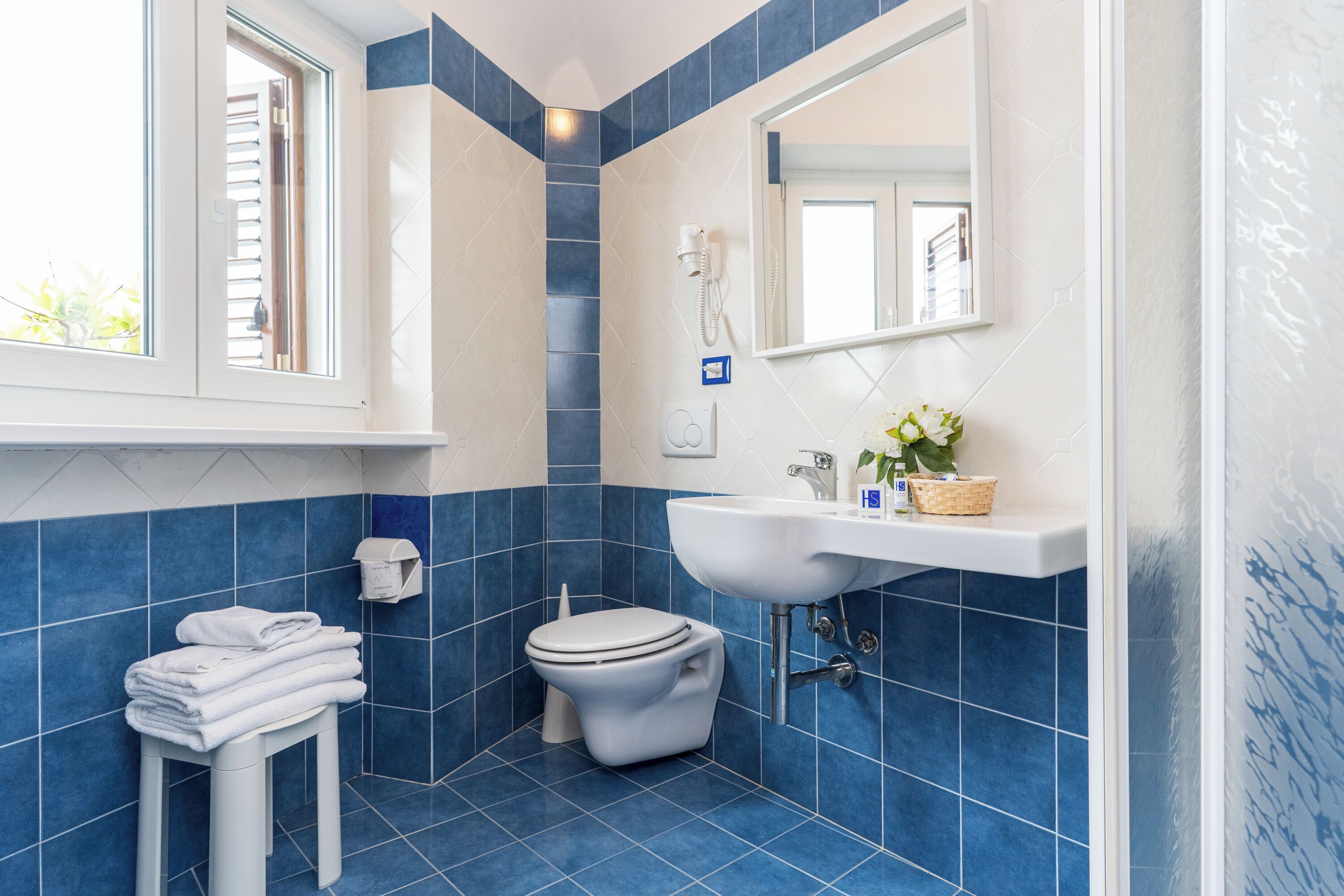 A bathroom with blue tiles, a white toilet, a sink, a mirror, and a window.