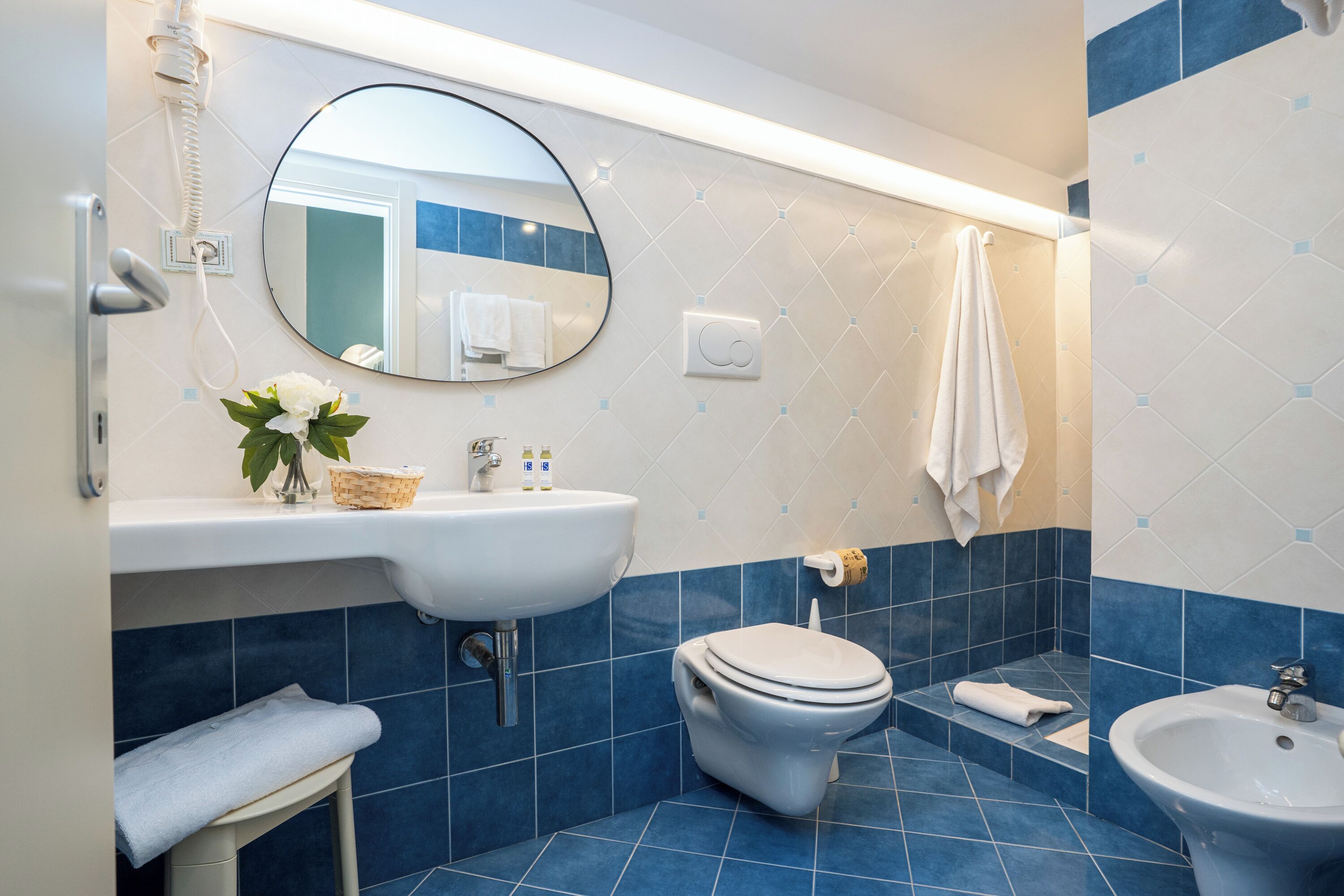 A bathroom with a white sink, a toilet, and a bidet. There is a mirror above the sink and a towel hanging on the wall.