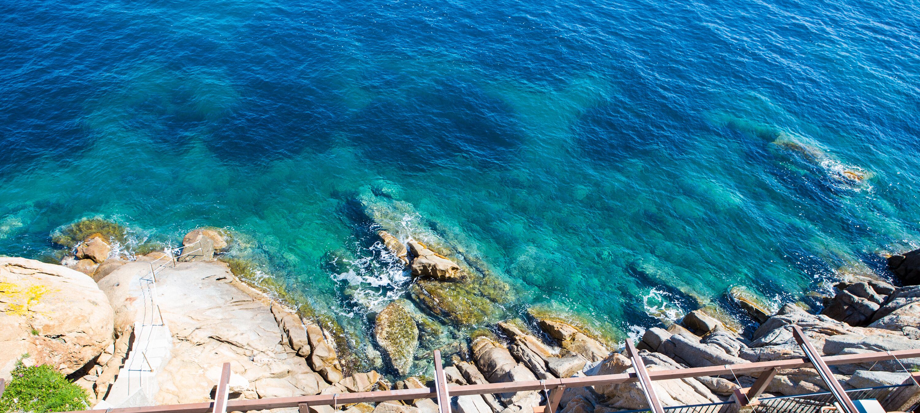 A coastal view with clear turquoise water, rocky shoreline, and a wooden staircase leading down to the water.