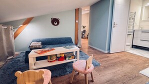 Une chambre à coucher aménagée en mezzanine, chaleureuse, avec un canapé bleu, une table de chevet blanche et deux fauteuils roses. On peut voir une décoration murale représentant un ours en bois, et une porte blanche donne accès à la salle de bain.