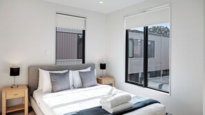 Une chambre moderne avec un grand lit, deux tables de chevet, une commode et une fenêtre donnant sur des immeubles.
