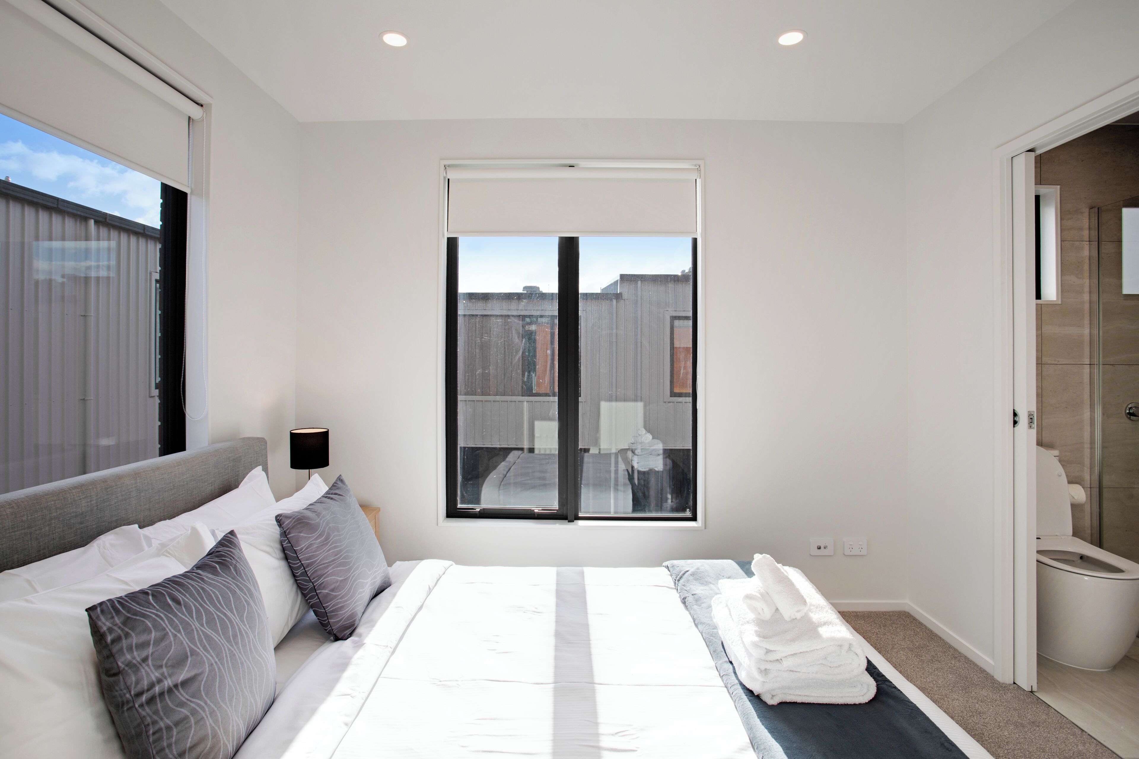 A modern bedroom with a large bed, a window with blinds, a bedside table with a lamp, and a view of the outside.