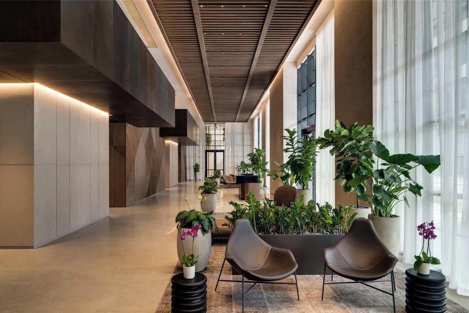 A spacious lobby with modern architecture, featuring a high ceiling, large windows, and a variety of potted plants.
