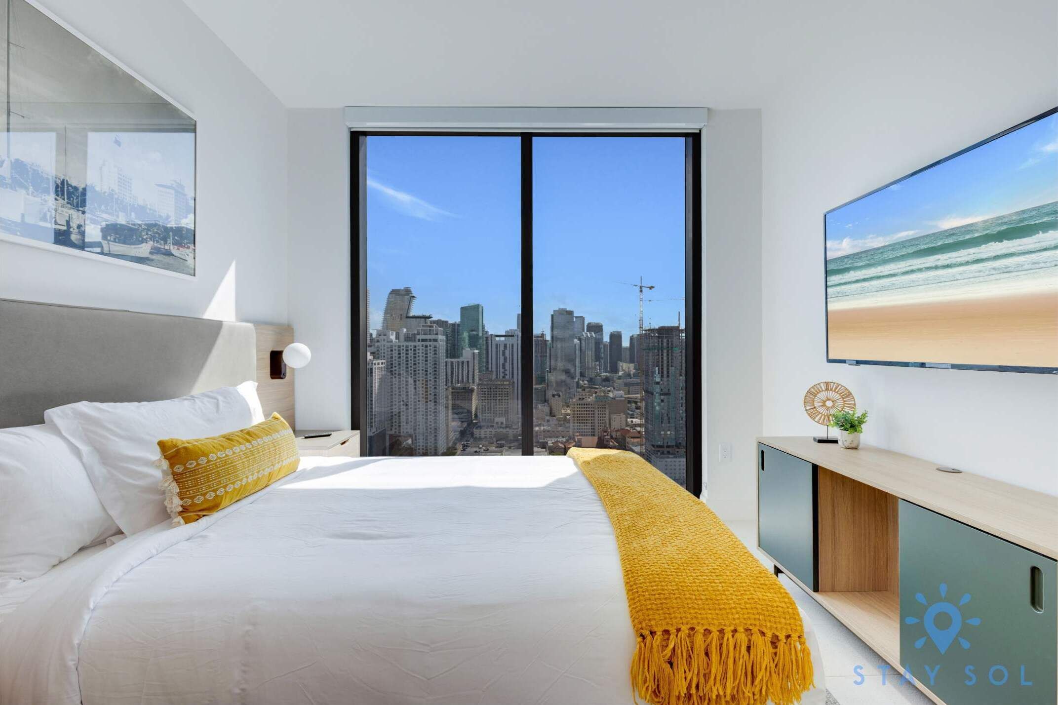 A modern hotel room with a large bed, a city view, a mounted TV, and a minimalist decor.