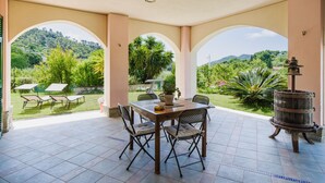 A covered patio with a table and chairs, a decorative metal water feature, and a view of a lush garden and hills in the background. - Casa Melograno by Interhome (Sestri Levante)