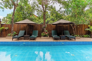 A swimming pool with four lounge chairs and two umbrellas, surrounded by a wooden fence and lush greenery. - Tiny Home (Siem Reap)