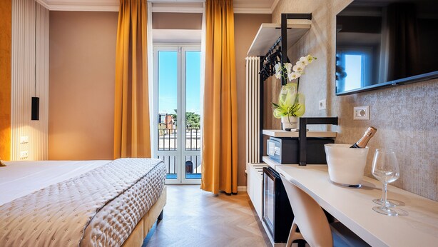 A hotel room with a bed, a desk with a TV, a minibar, and a view of the outdoors. - Colonna Inn (Rome)
