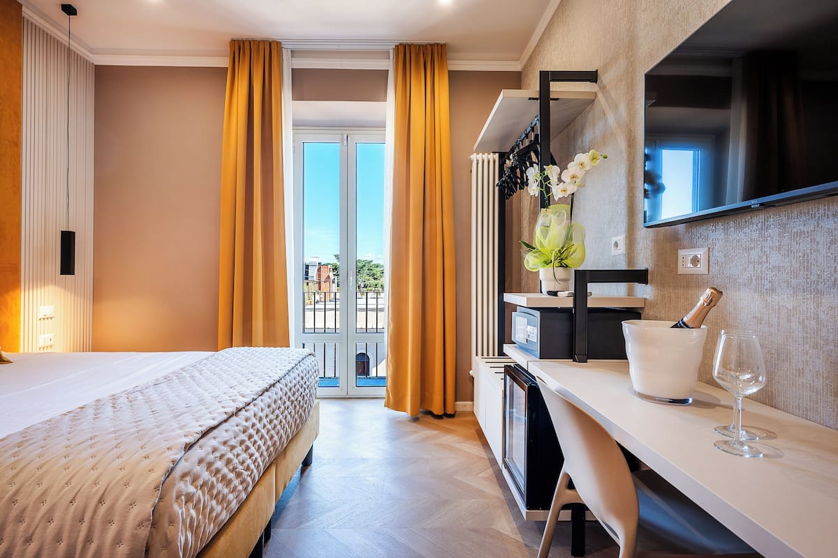 A hotel room with a bed, a desk with a TV, a minibar, and a view of the outdoors.