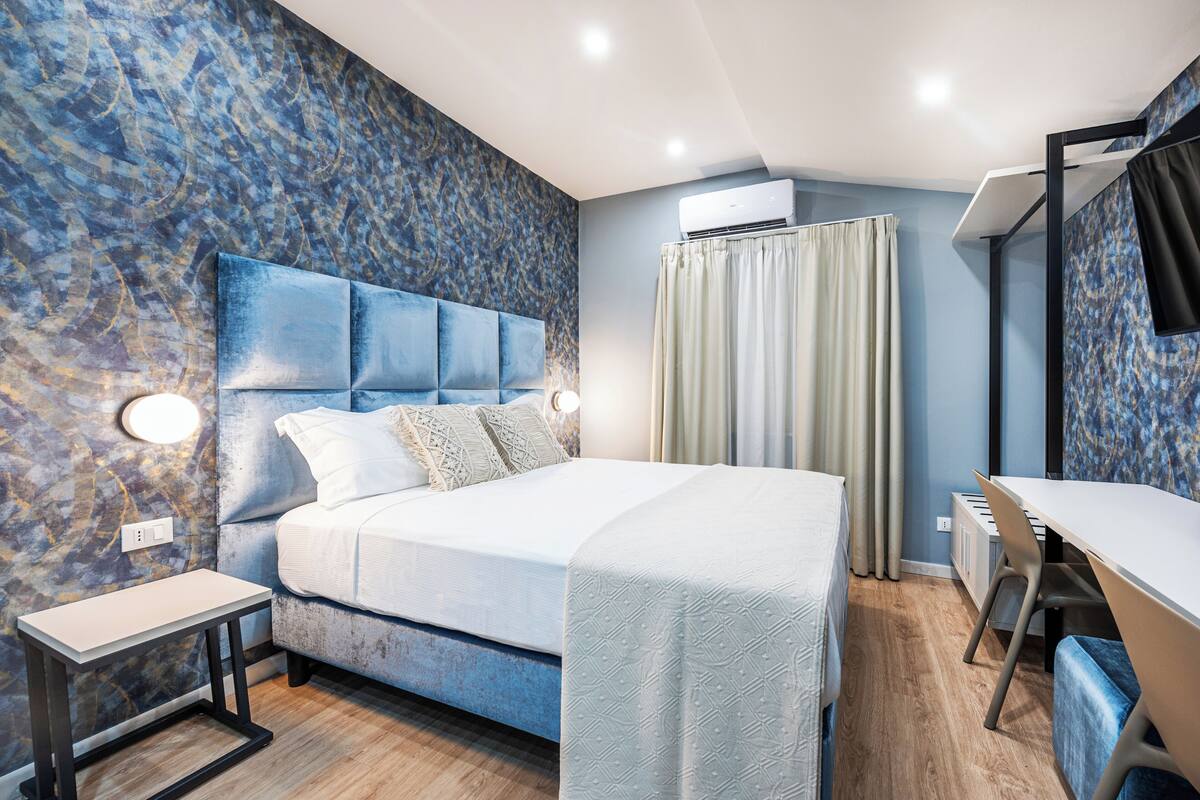 A modern bedroom with a large bed, bedside tables, a desk, and an air conditioning unit.