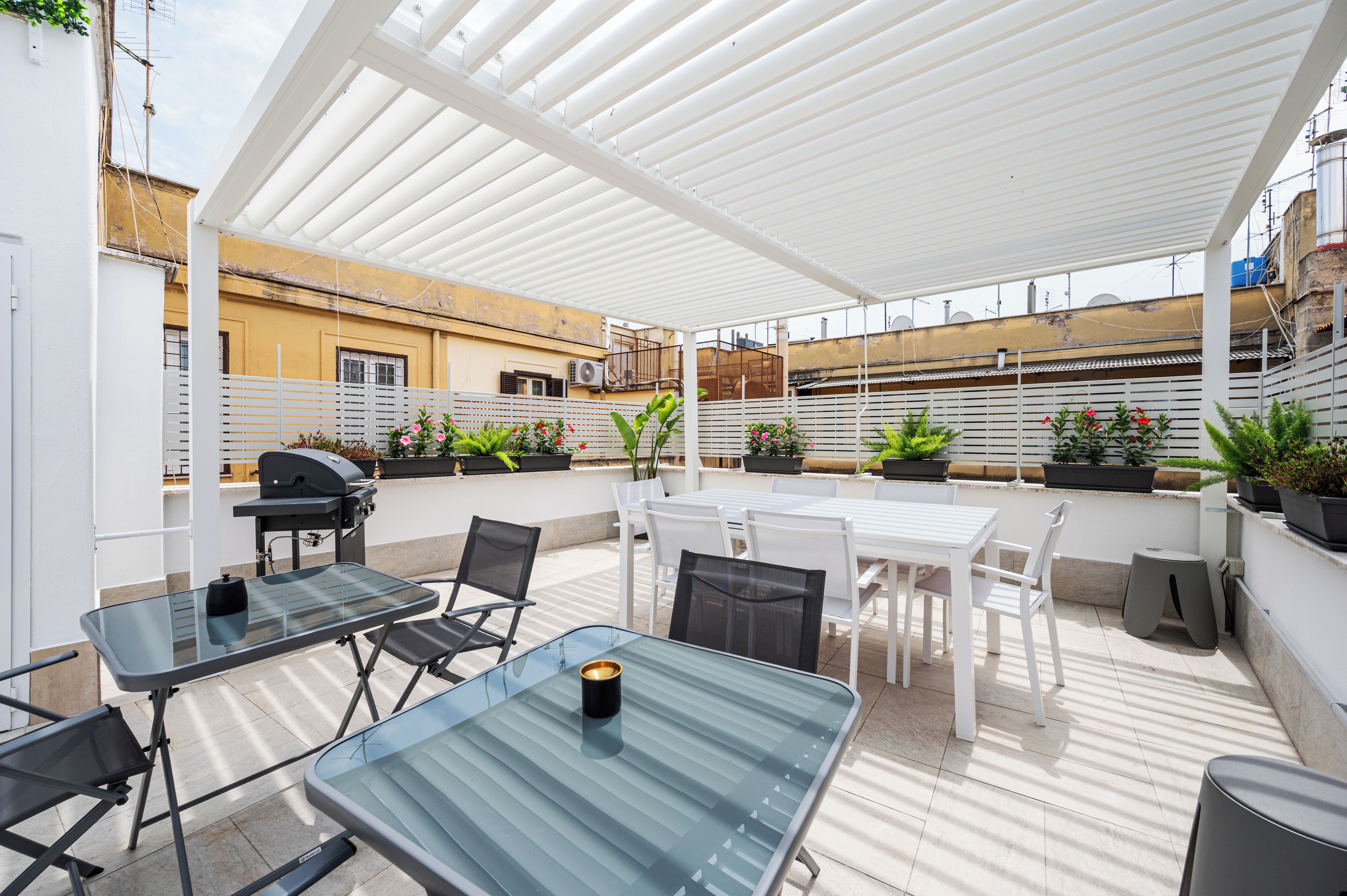 Une terrasse extérieure dotée d’une pergola blanche, de tables en verre et de chaises. On y trouve également des plantes en pot et un barbecue.