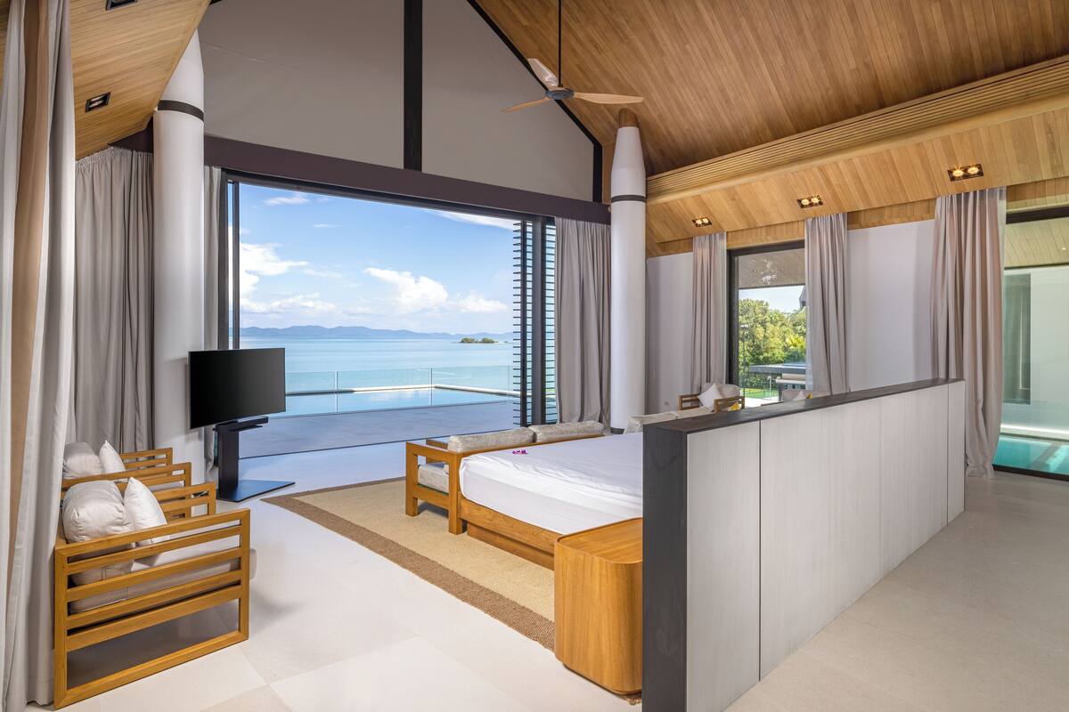 A modern bedroom with a large bed, a TV, and a balcony with a view of the sea.