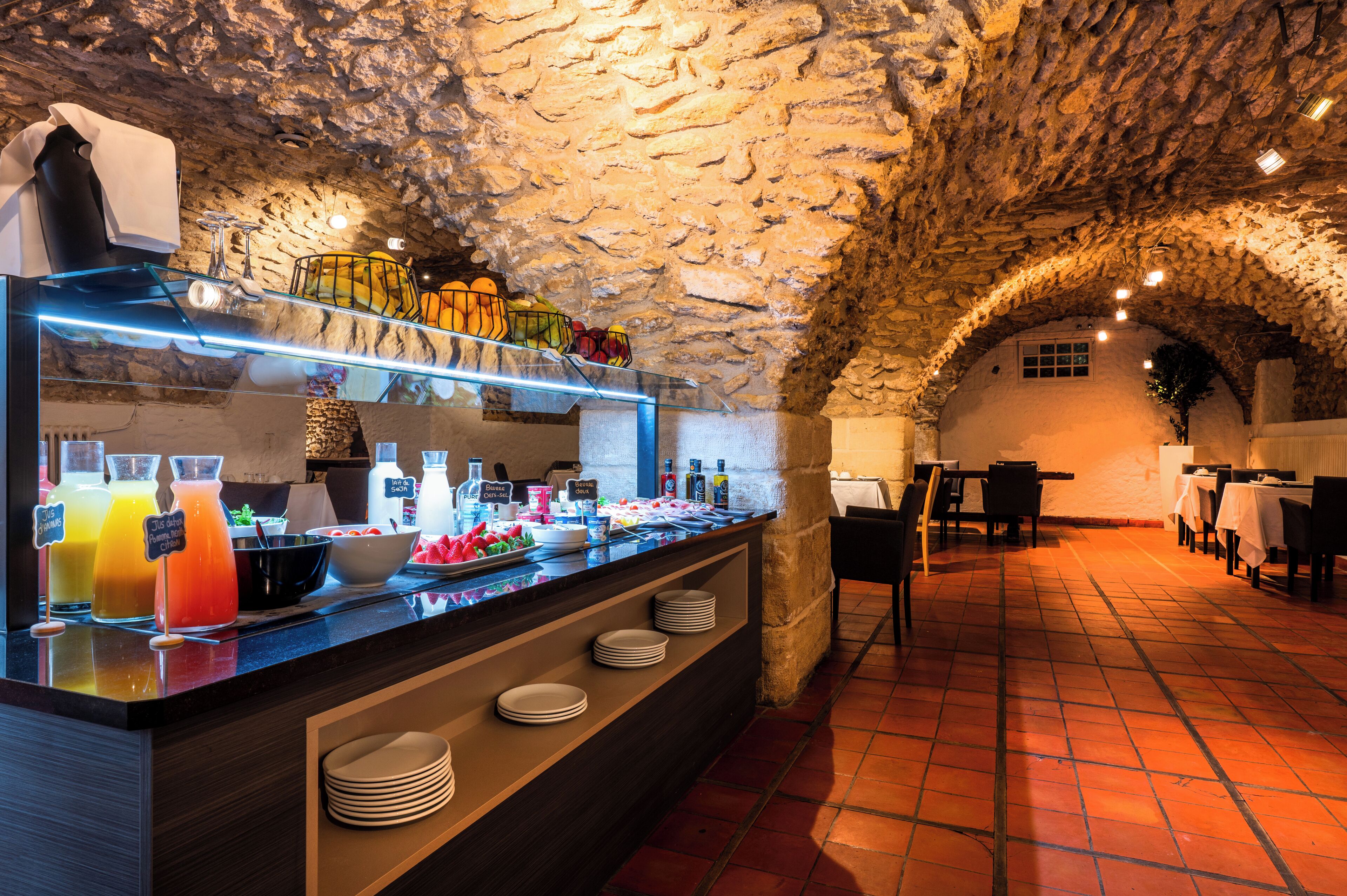 Un bar à petit-déjeuner aux murs en pierre, proposant une variété de plats et de boissons, avec un sol carrelé et des tables et des chaises en arrière-plan.