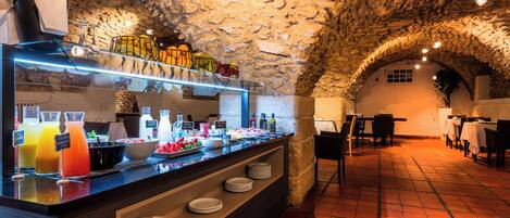 Un bar à petit-déjeuner aux murs en pierre, proposant une variété de plats et de boissons, avec un sol carrelé et des tables et des chaises en arrière-plan.