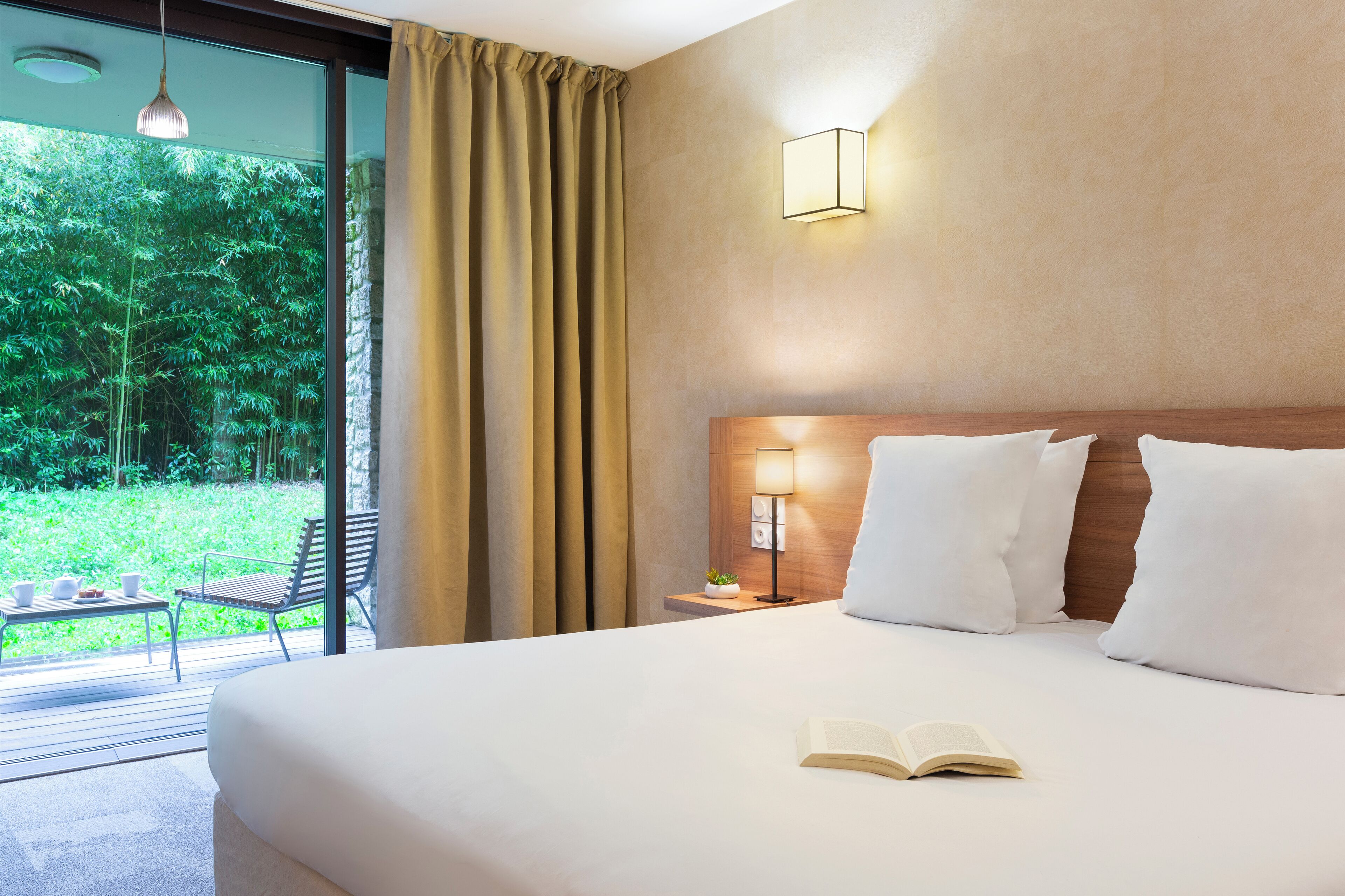 Een hotelkamer met een groot bed, een nachtkastje met lamp, een raam met gordijnen en uitzicht op groen.