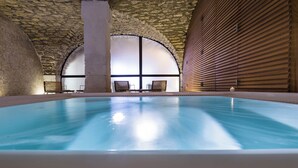 Una piscina interna con pareti in pietra e pannelli di legno, panche e un'ampia finestra ad arco.