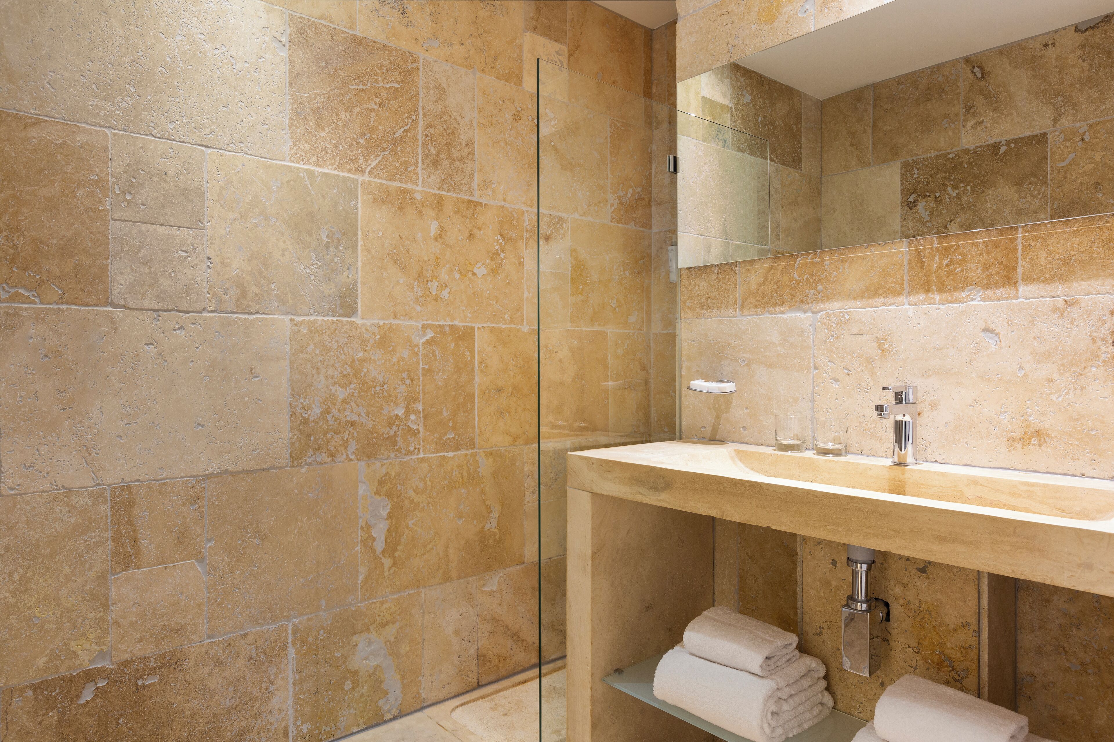 A modern bathroom with beige stone tiles, a glass shower enclosure, a wooden vanity with a sink, and a mirror.
