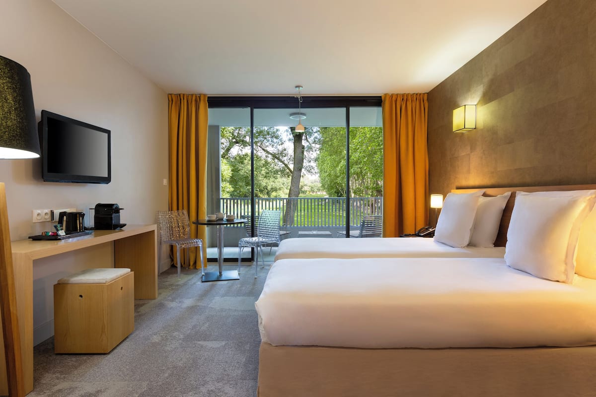 Habitación de hotel con dos camas, un escritorio, televisor y balcón con vistas a árboles.