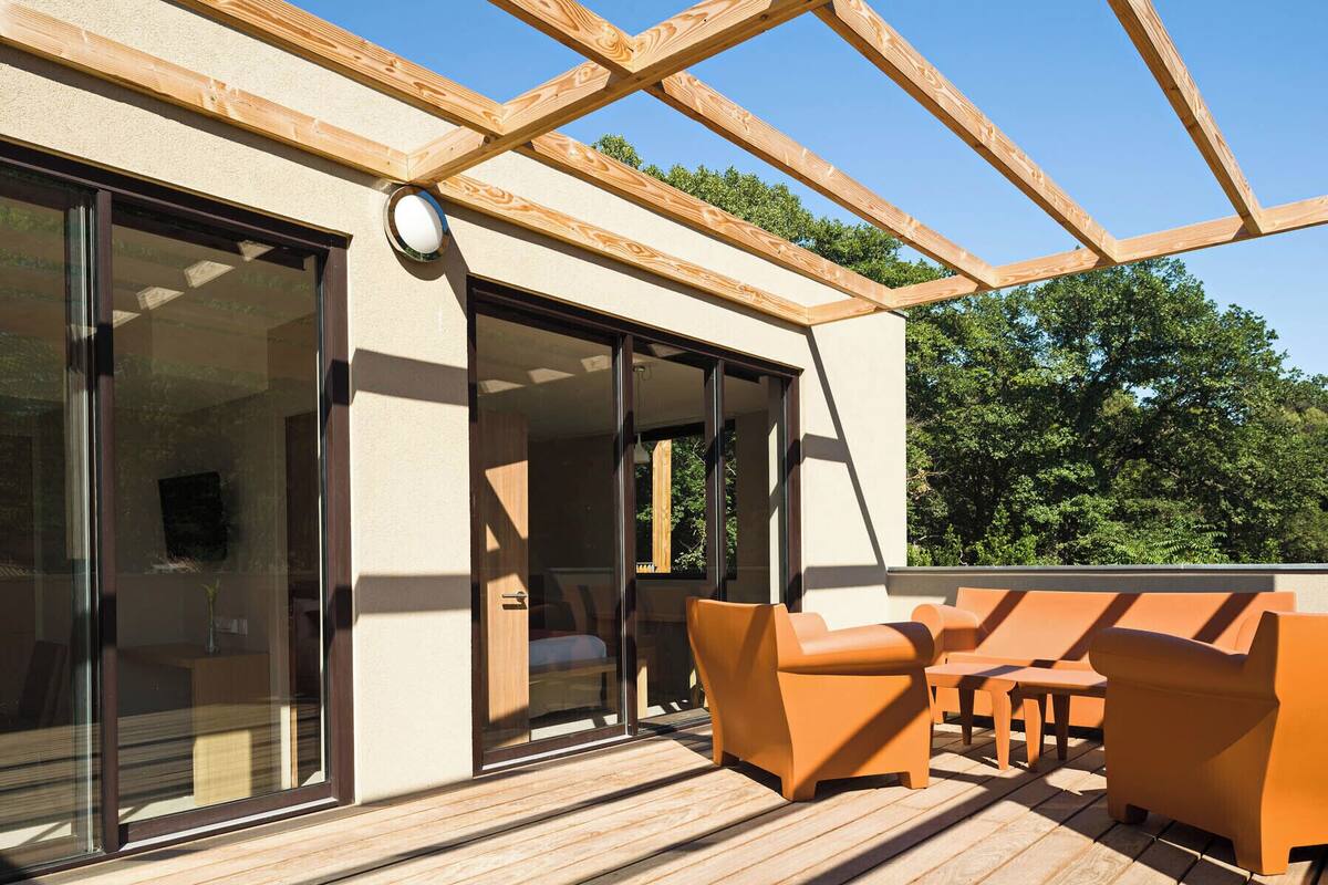 Una terraza de madera con mobiliario exterior, puertas correderas de vidrio grandes y una vista despejada de los árboles circundantes.