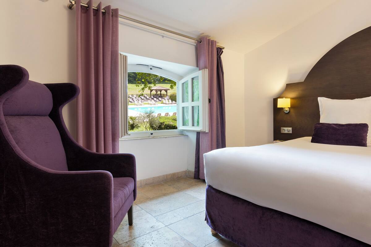 Habitación de hotel con cama, una silla, ventana con cortinas y vista a una piscina y sillas de descanso.