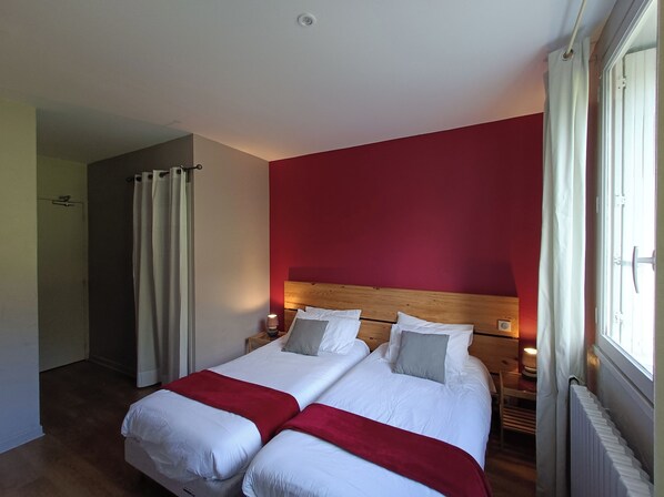 A double bed with white bedding and a red throw, a wooden headboard, a nightstand with a lamp, and a window with curtains. - CONTACT HOTEL LE BOIS SAUVAGE (Saint-Agrève)