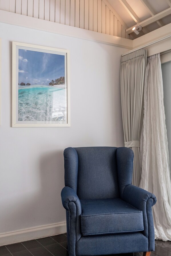 A room with a blue armchair, a framed picture of a beach scene, and a white curtain.