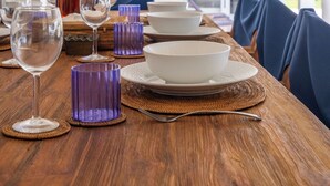 Mesa de comedor dispuesta con copas, fuentes y una botella decorativa, sillas y vistas al exterior.
