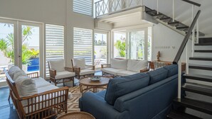 Un amplio salón con escalera, mobiliario de mimbre y ventanas grandes.