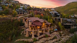 A large, stone-clad house with multiple levels and a prominent wrap-around porch, surrounded by a mountainous landscape.