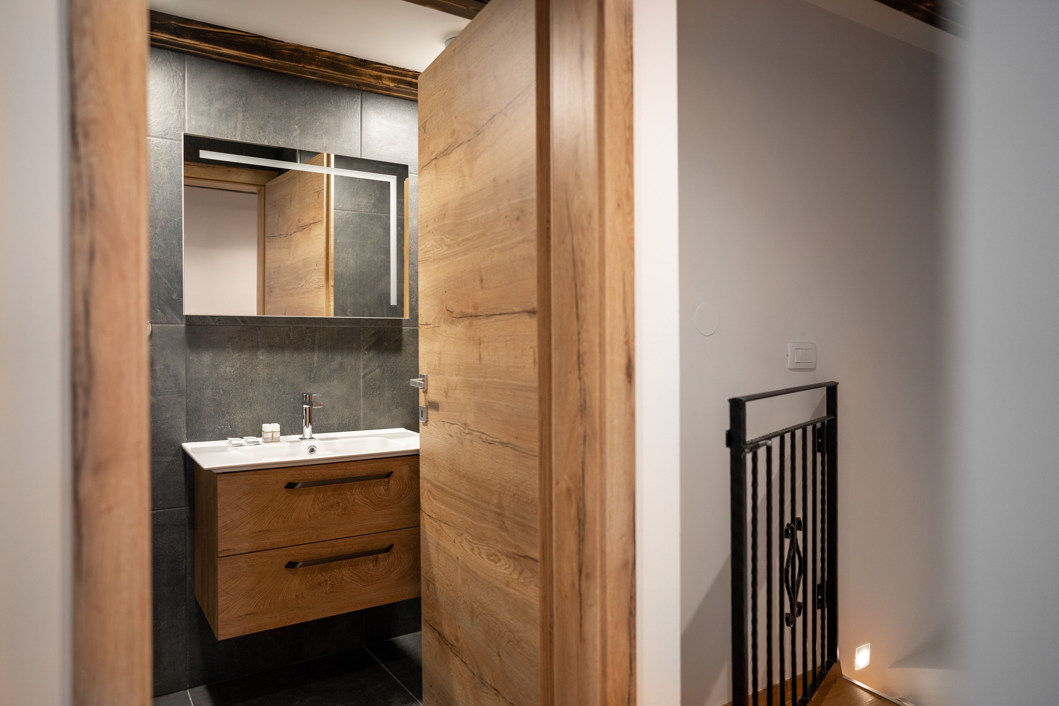 Un bagno moderno con un mobile vanitario in legno, uno specchio e una parete rivestita di piastrelle nere.