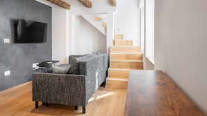 A modern living room with a grey sofa, a wooden coffee table, and a flat-screen TV mounted on a dark wall. A staircase with wooden steps leads to an upper floor. - Blue Sky Duplex (Piran)