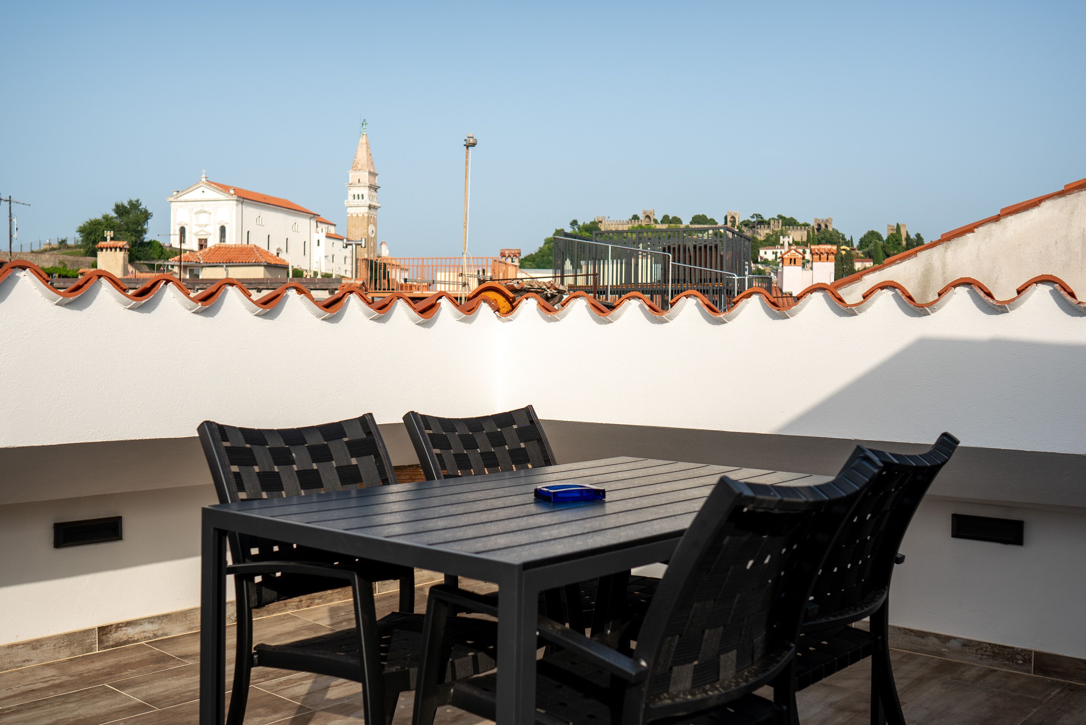 Een terras op het dak met een tafel en stoelen, een helder uitzicht op een historisch gebouw met een klokkentoren, en een stadsgezicht op de achtergrond.