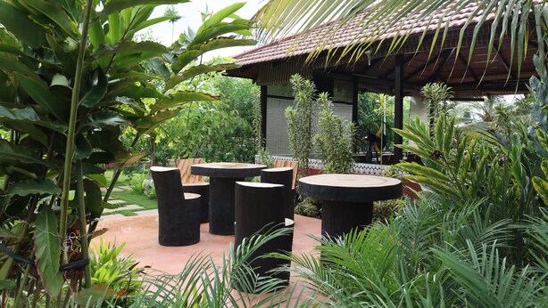 An outdoor seating area with round tables and chairs, surrounded by lush greenery and a traditional-style building with a tiled roof. - Abhishta Agri Farm Stay (Kunigal)